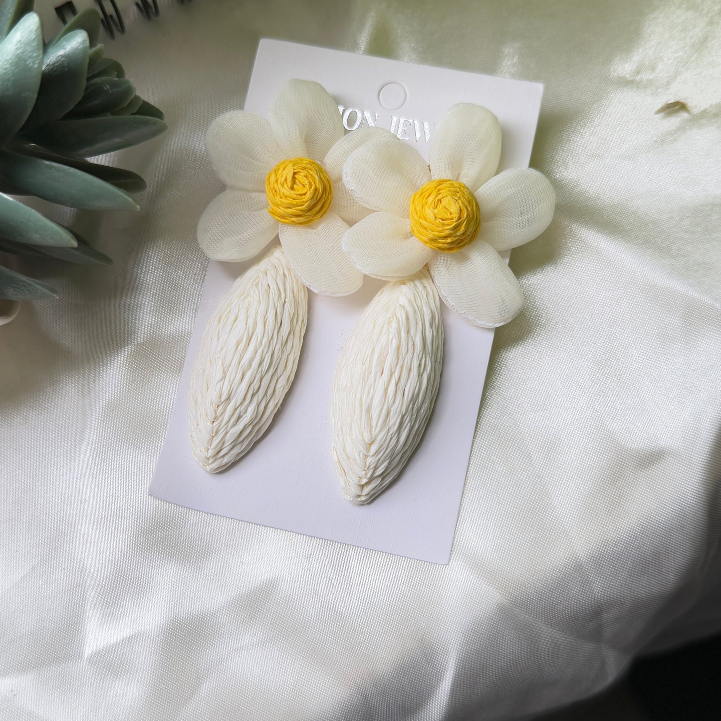 Pair of white floral earrings with yellow centers on a white card, placed on a light fabric background.