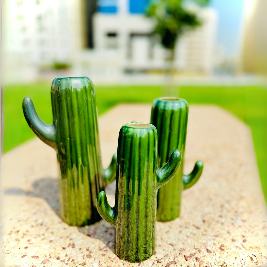 Three green cactus-shaped objects on a sandy surface with a blurred outdoor background.