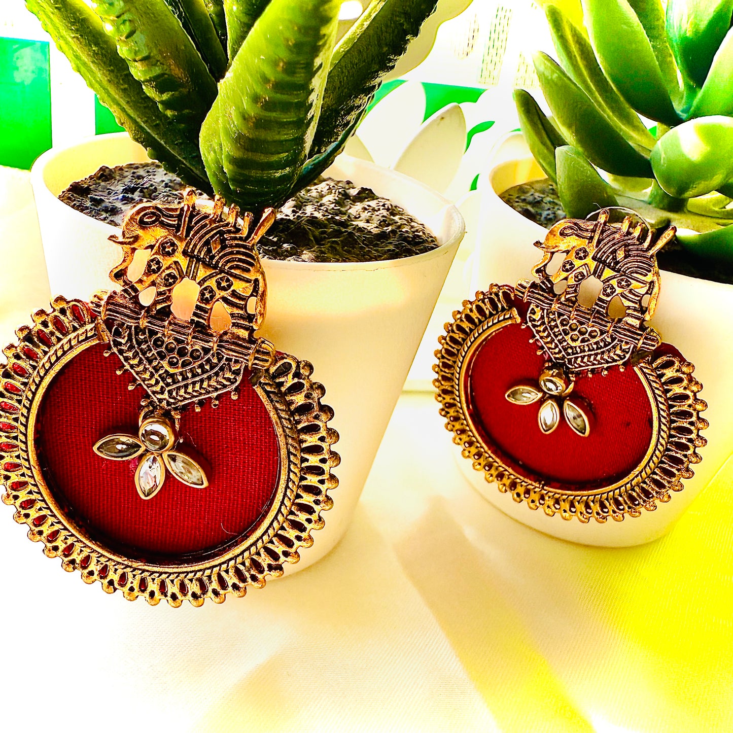 Ornate earrings with red center and gold detailing on a white surface with green plants in the background.
