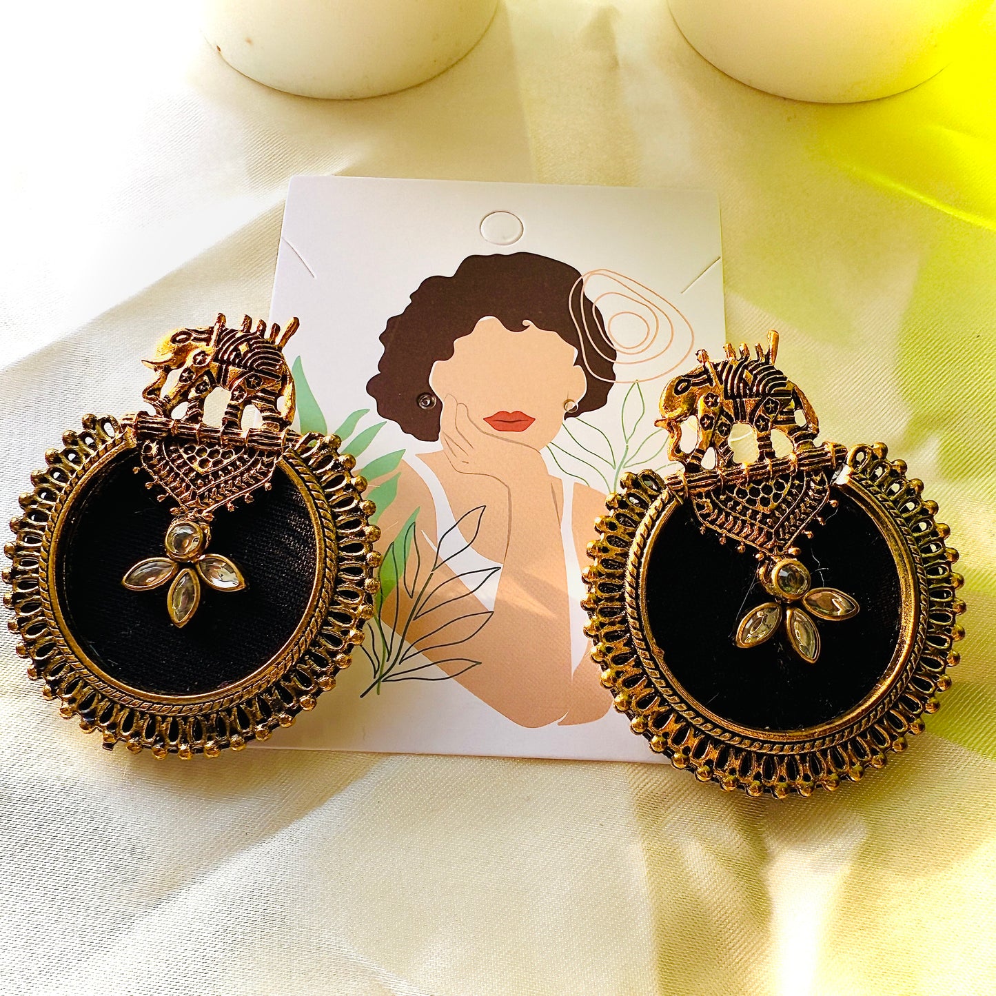 Ornate earrings with black center and gold detailing on a white card with a woman's face.