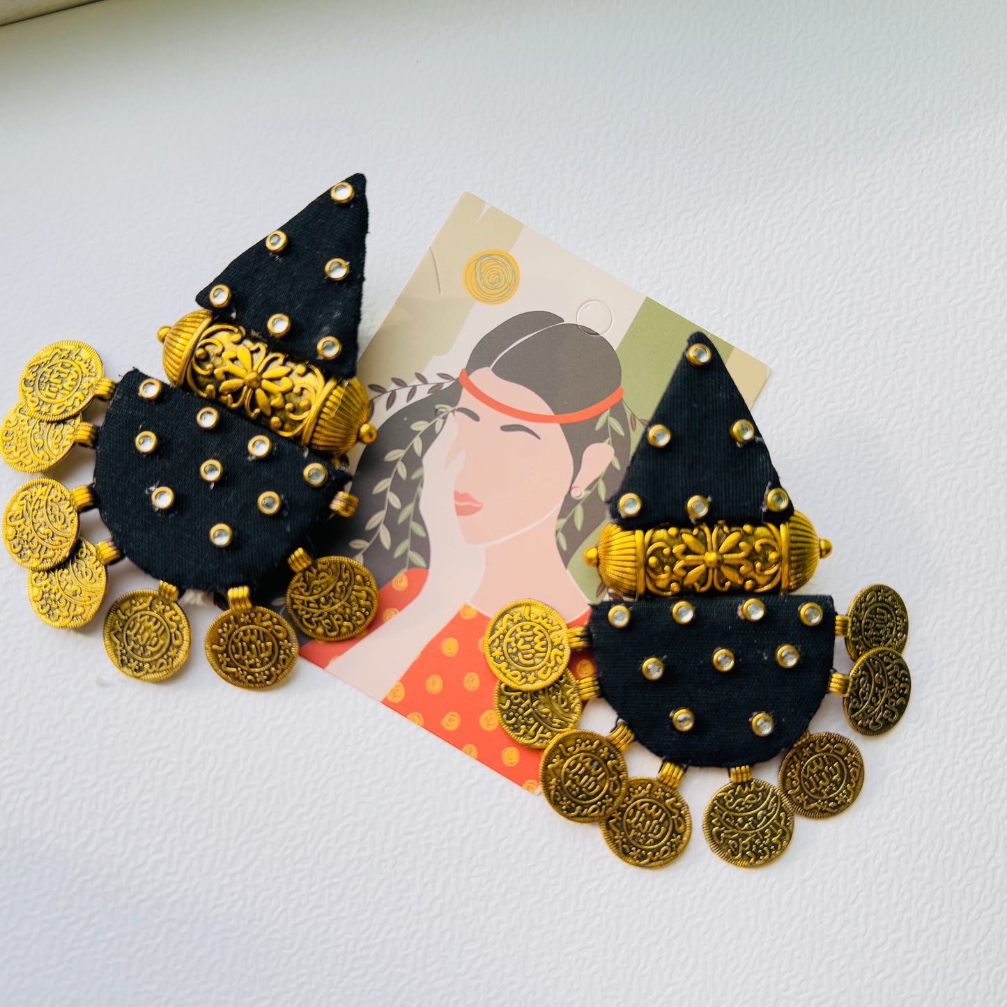 Black and gold earrings on a white background with a decorative card featuring a woman's face.
