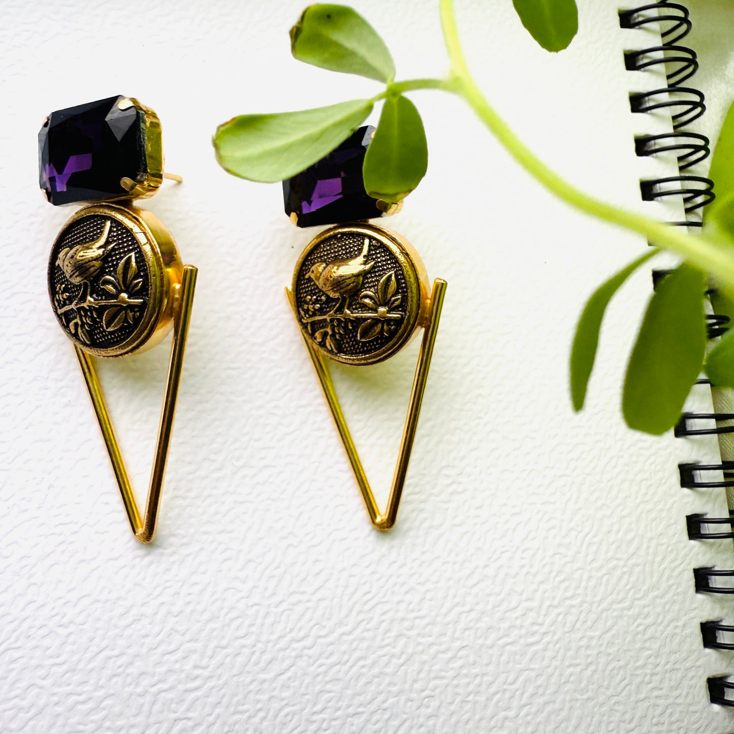 Gold earrings with purple gemstones on a white surface with a plant and notebook.
