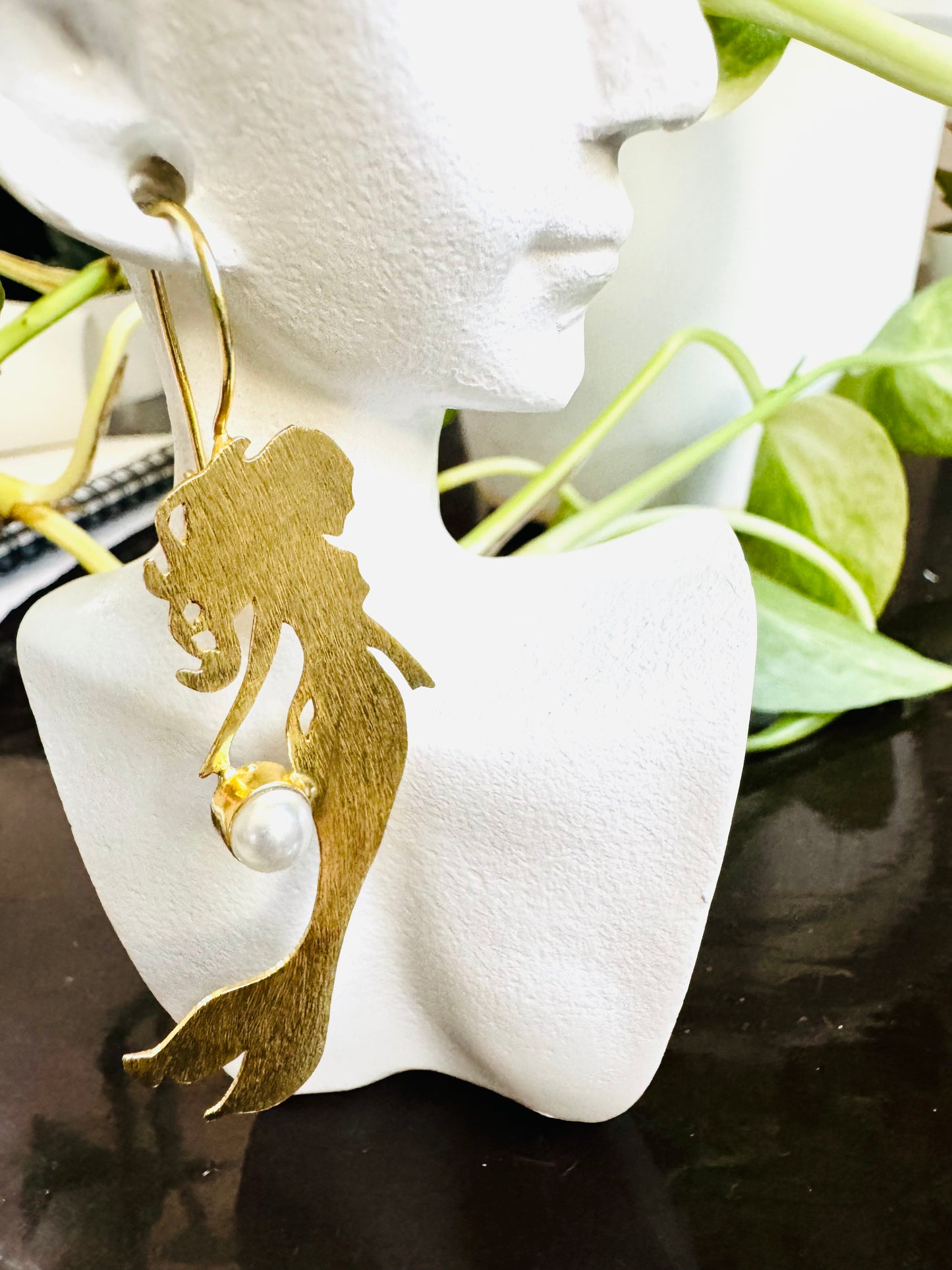Gold earrings with a pearl on a white stand with green leaves in the background