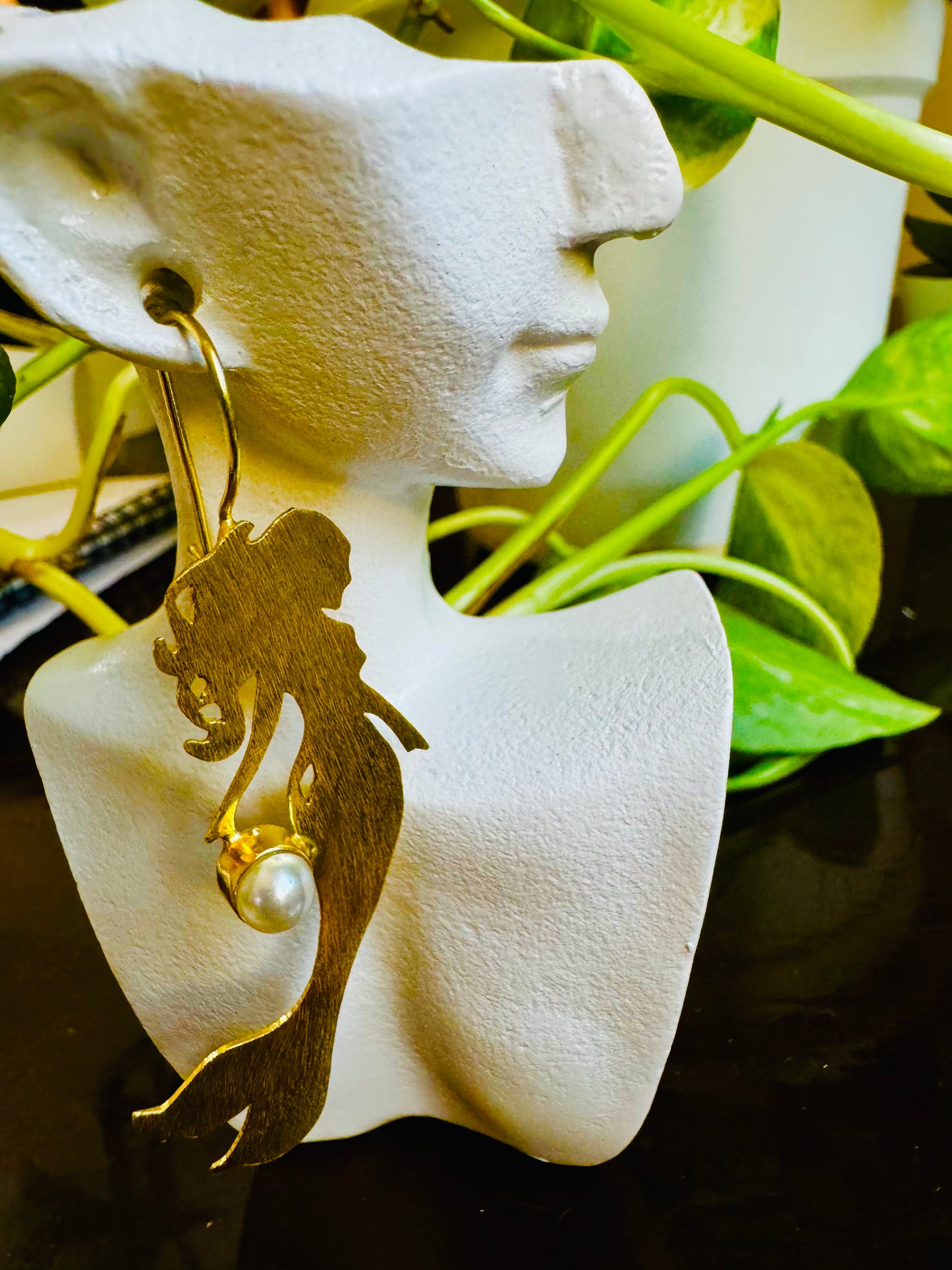 Gold earrings with a pearl on a white earring stand with green leaves in the background