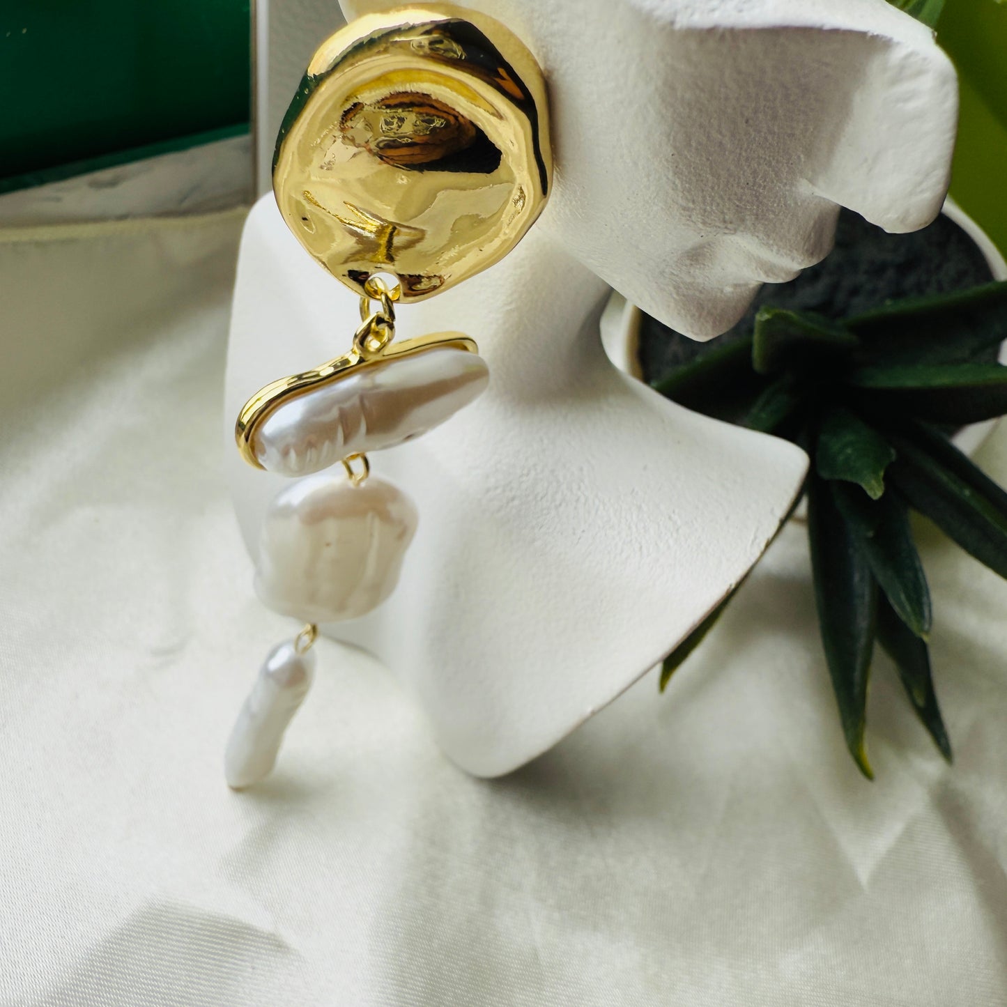 Gold and white earrings on a mannequin ear with a green plant in the background