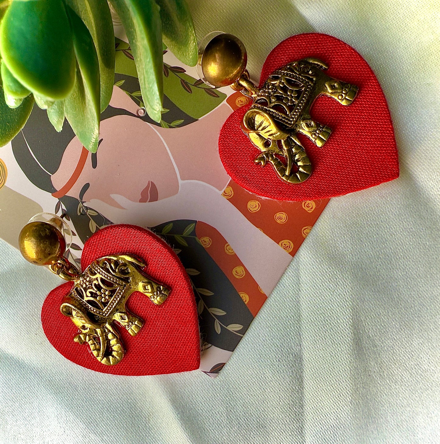 Heart-shaped red fabric clips with gold turtle designs on a white surface with a plant in the corner.