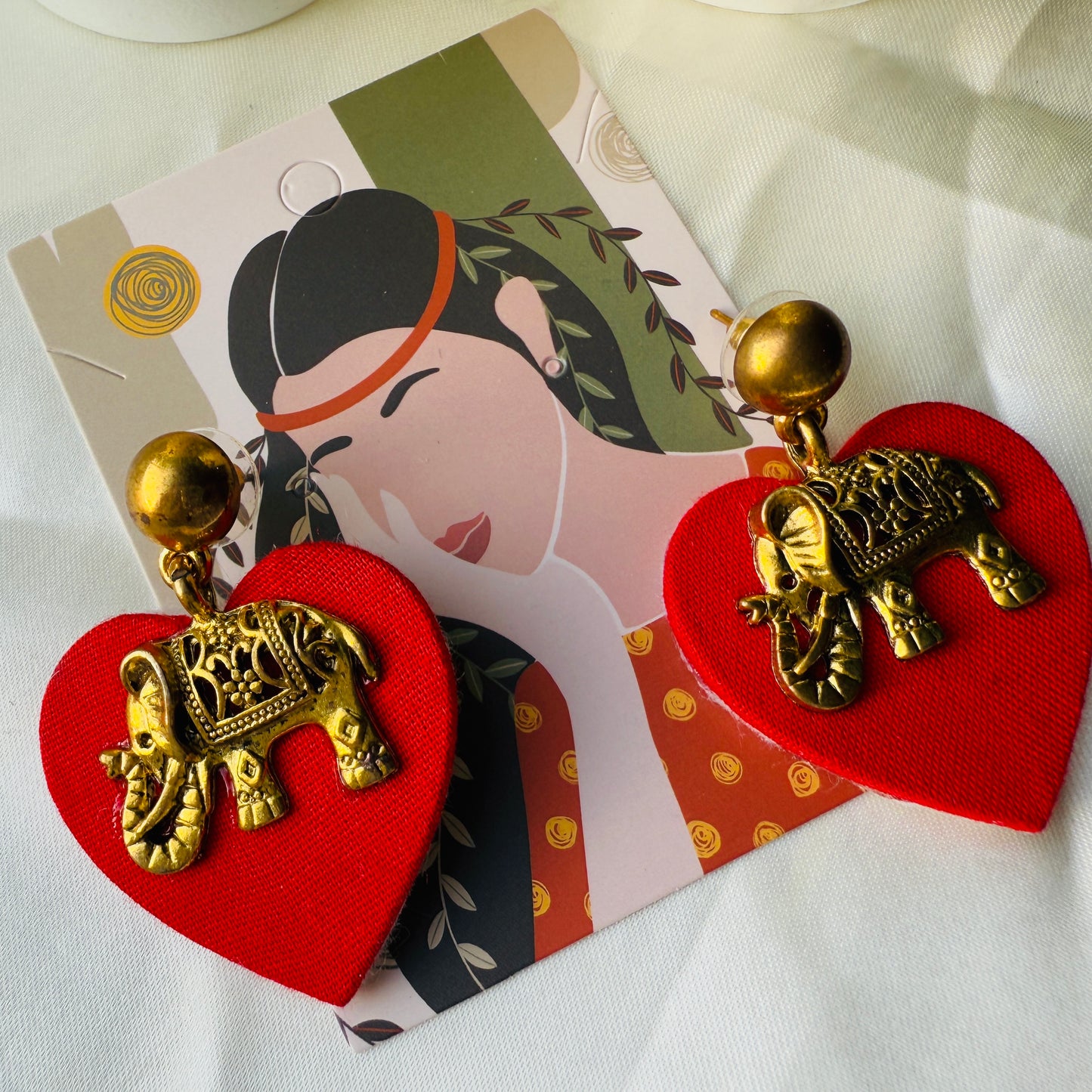 Red heart-shaped earrings with gold elephant designs on a decorative card.