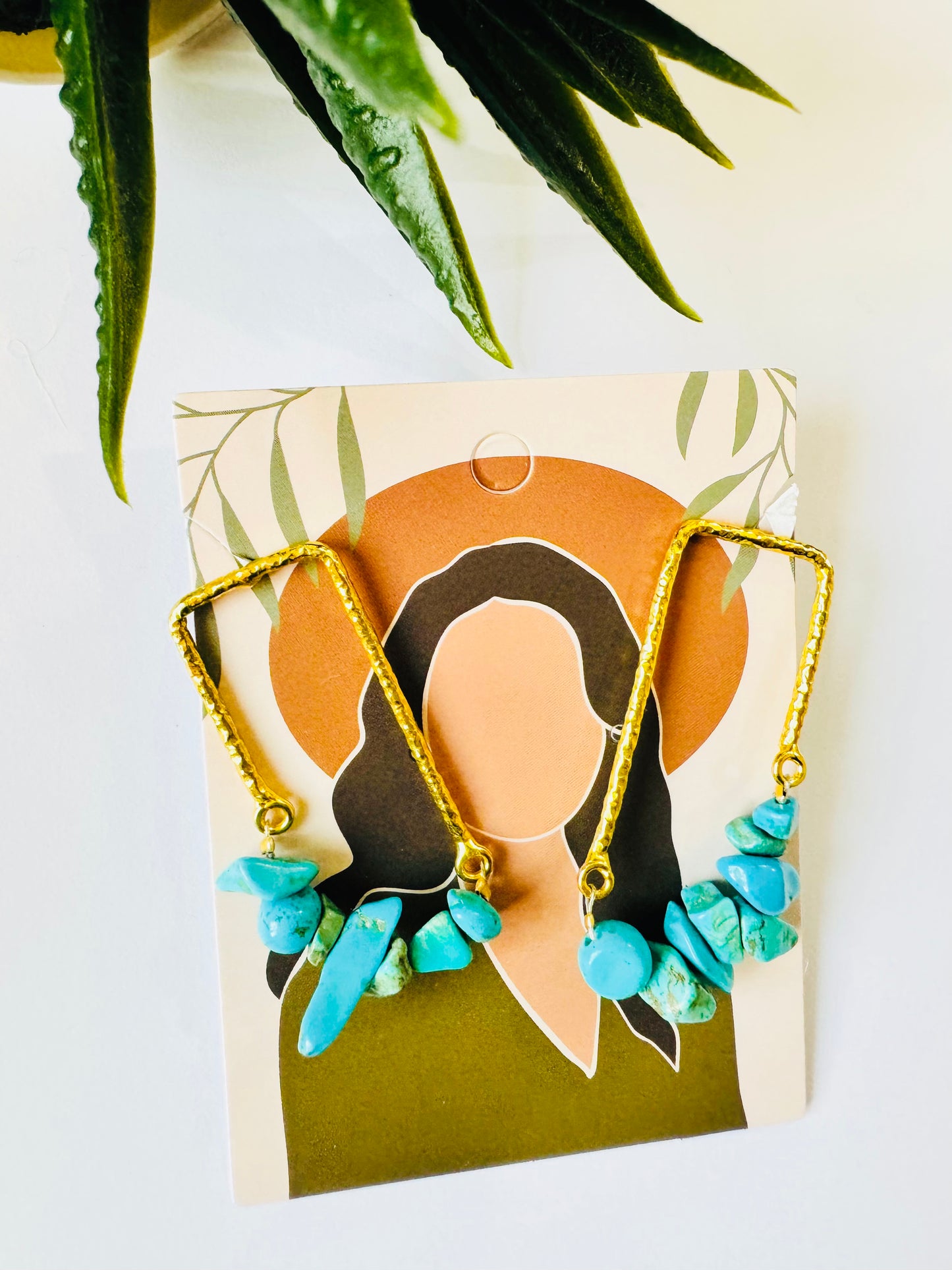 Gold earrings with turquoise stones on a decorative card with a plant in the background