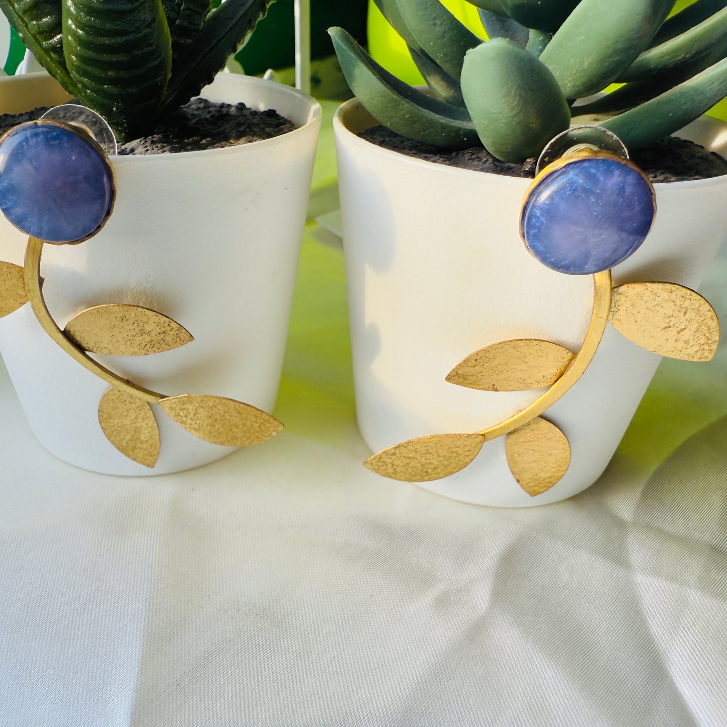 Pair of earrings with gold leaf design and blue gemstone in white pots with plants.