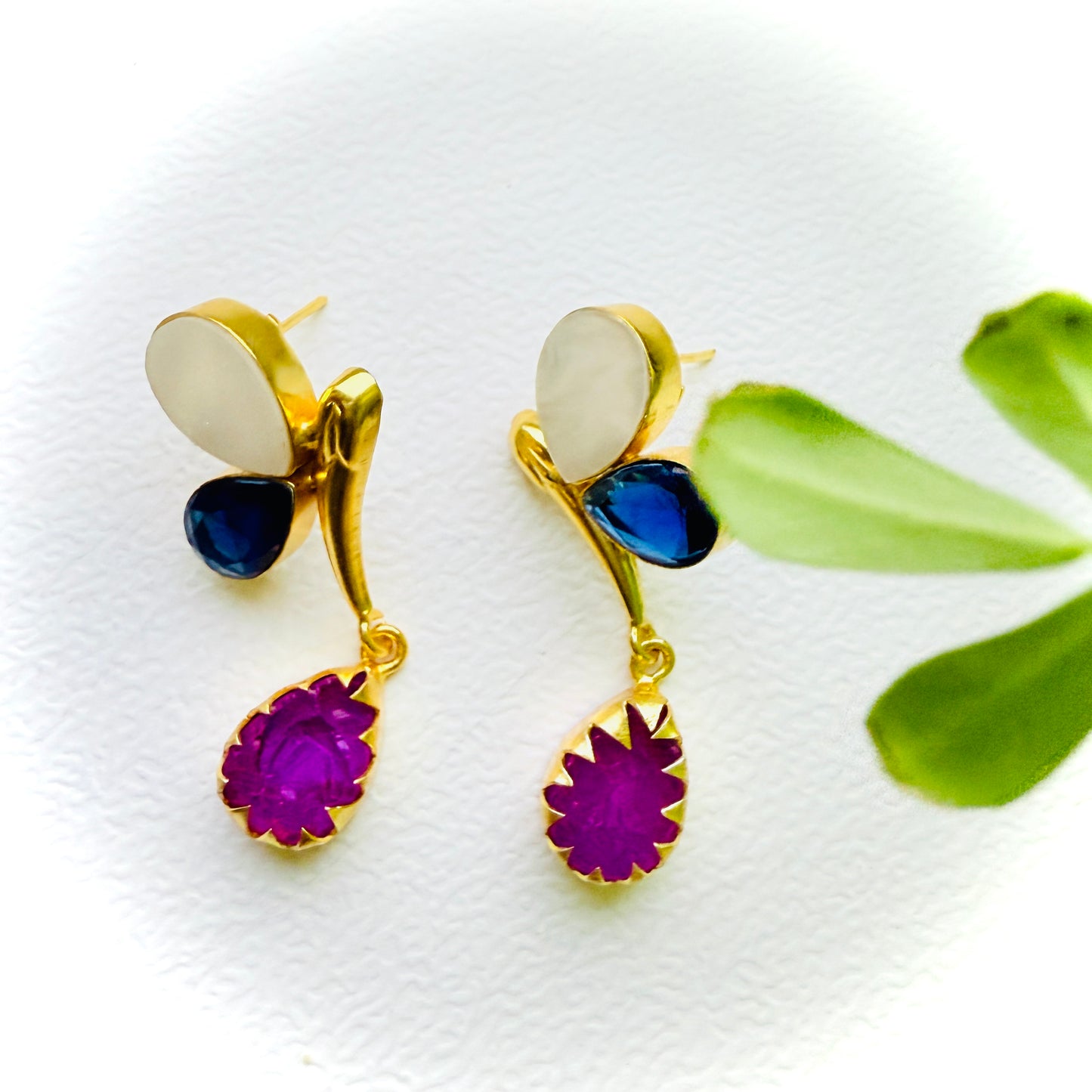 Gold earrings with purple and blue stones on a white background with green leaves.