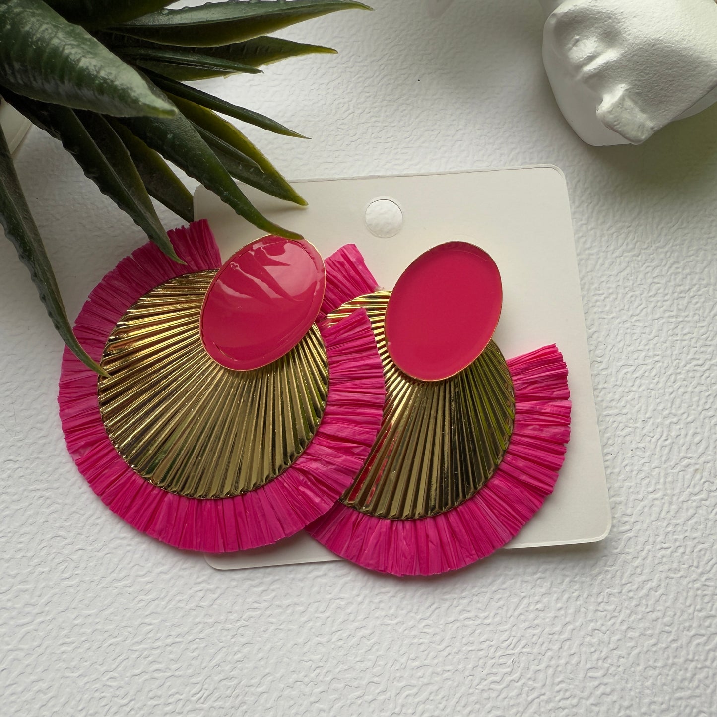 Pink and gold earrings on a white card with a plant in the background