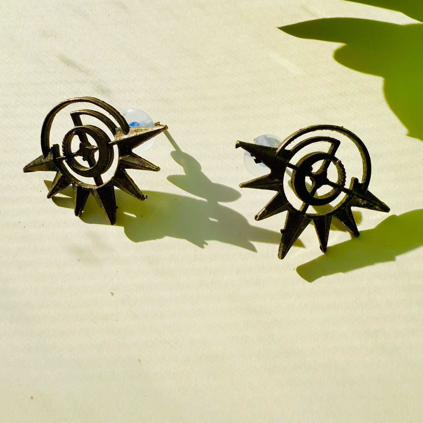 Pair of black and gold earrings on a beige surface with a green leaf shadow.