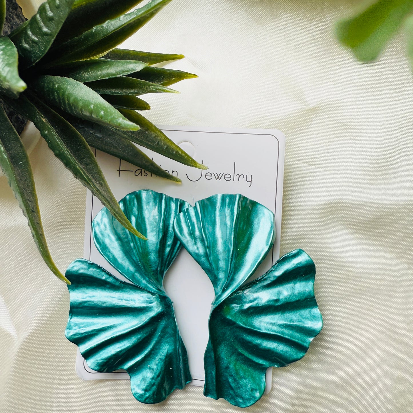 Teal leaf-shaped earrings on a white card with 'Fashion Jewelry' text, placed on a light fabric background with green leaves.