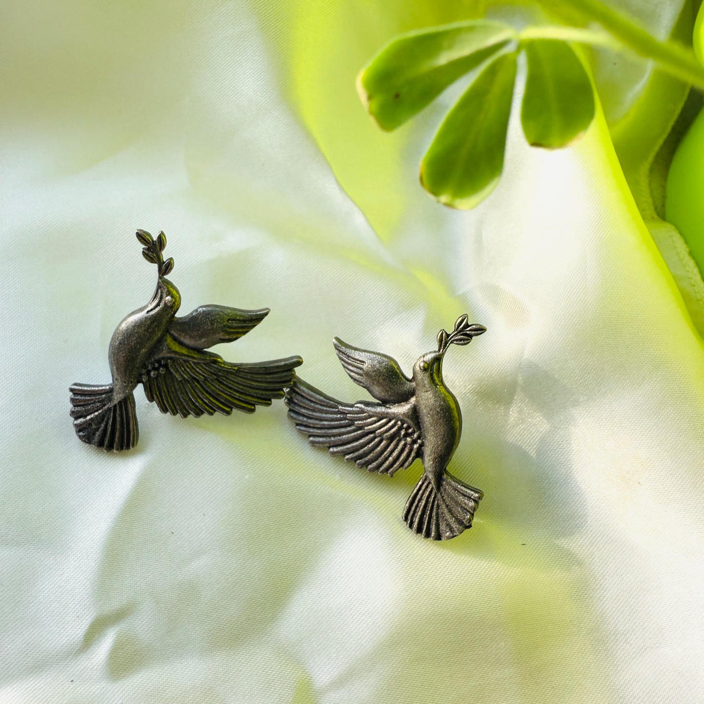 Bronze bird earrings on a white fabric background with green leaves.