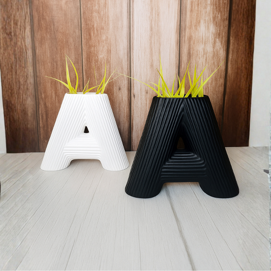 Two letter 'A' shaped planters, one white and one black, with grass-like plants on a wooden floor.