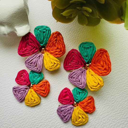 Colorful flower-shaped earrings on a textured white surface with green leaves in the background.