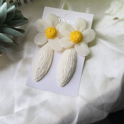 Pair of white floral earrings with yellow centers on a white card, placed on a light fabric background.