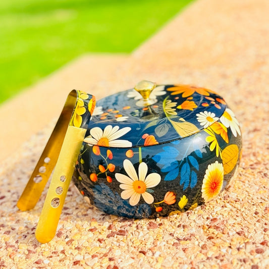 Decorative container with floral design and a yellow tool on a sandy surface