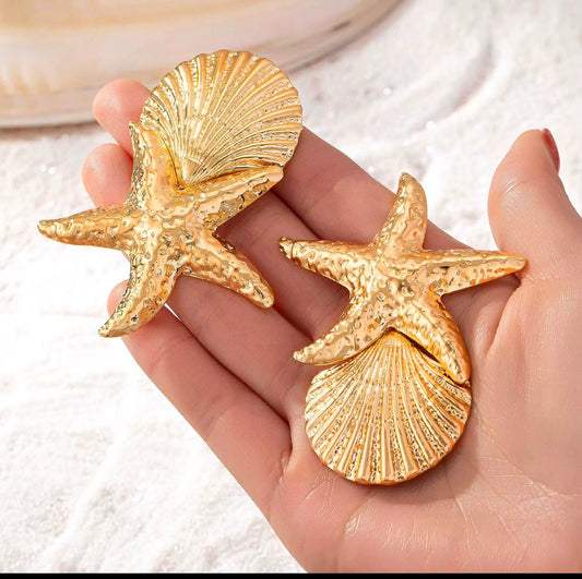 Gold starfish and shell earrings held in a hand against a white background
