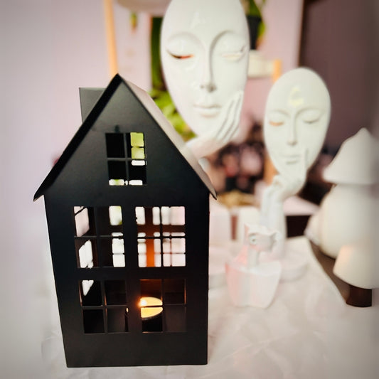 Black house-shaped lantern with a lit candle inside, placed on a surface with decorative items in the background.