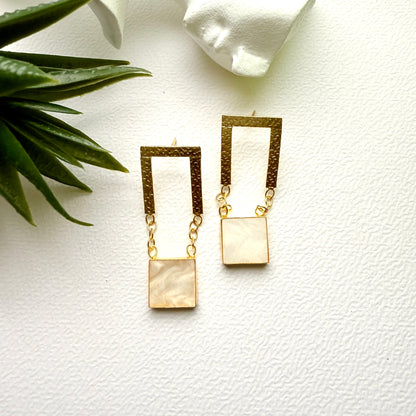 Gold earrings with square pendants on a white surface with a plant leaf.