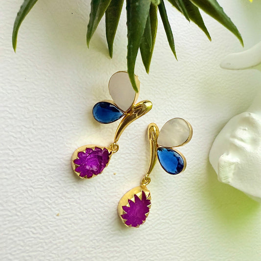 Gold earrings with purple and blue stones on a white surface with green leaves in the background